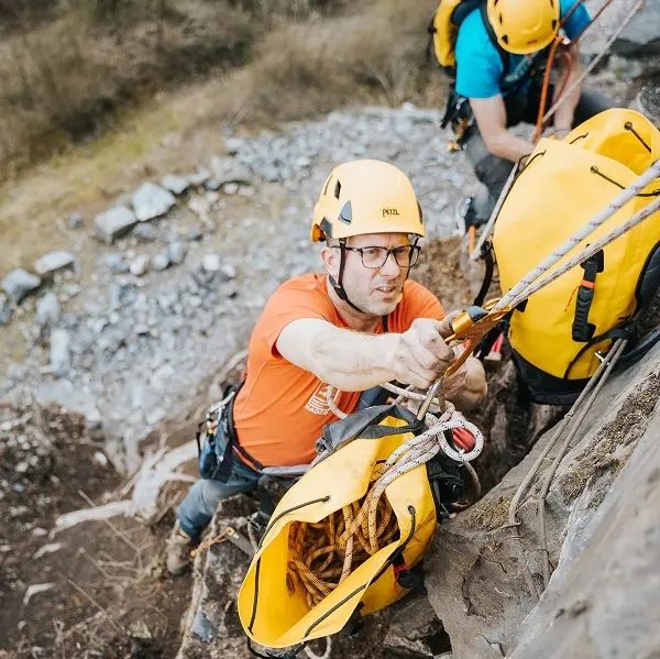 Twee mensen met helmen en klimgordels beklimmen een rotsachtige helling. De persoon vooraan, met een bril en een oranje shirt, houdt zich vast aan een touw en draagt een gele tas vol klimuitrusting. Achter hem is nog een klimmer te zien. Twee mensen met helmen en klimgordels beklimmen een rotsachtige helling. De persoon vooraan, met een bril en een oranje shirt, houdt zich vast aan een touw en draagt een gele tas vol klimuitrusting. Achter hem is nog een klimmer te zien.